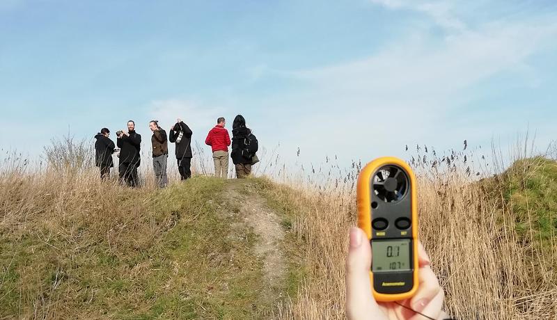 Een student voert metingen uit met een klein apparaatje. Op de achtergrond staan studenten op een verhoging in het landschap.