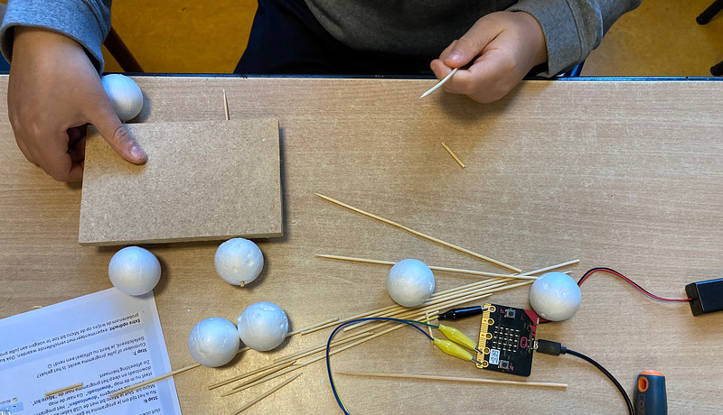 Foto van kinderhanden bij een tafel. Op de tafel liggen allerlei voorwerpen, zoals satéprikkers, piepschuim, kabels en een schroevendraaier.