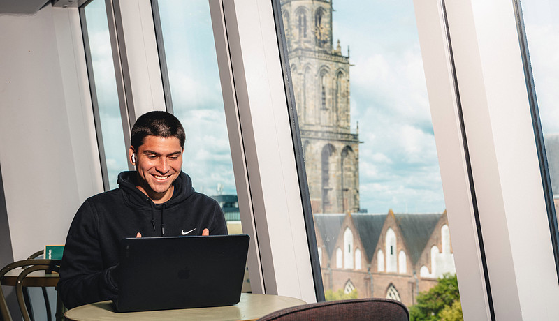 Student smiling while studying on a laptop in a modern building with large slanted windows, with the Martini Tower of Groningen visible in the background.