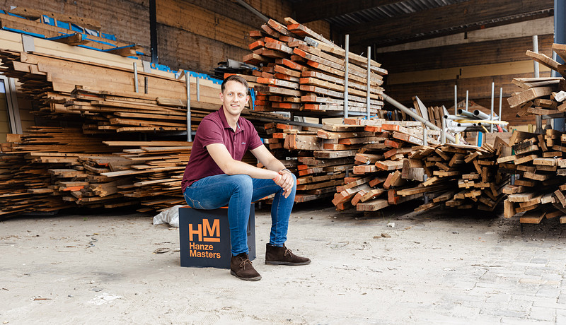 Alumnus Koen Rooseboom sitting on Master Up box at work with pile of wood boards behind him