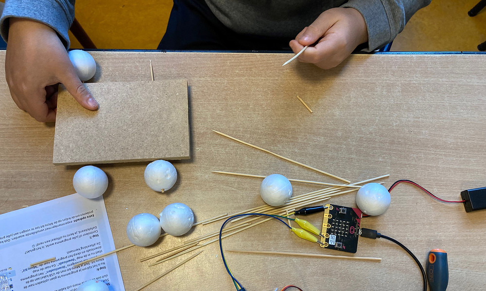 Foto van kinderhanden bij een tafel. Op de tafel liggen allerlei voorwerpen, zoals satéprikkers, piepschuim, kabels en een schroevendraaier.