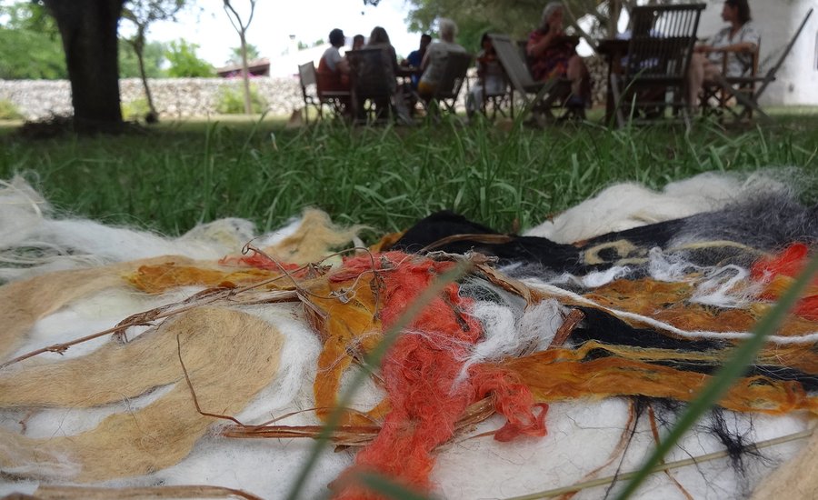 Mensen zitten op stoelen in een grasveld. Op de voorgrond liggen gekleuren plukken wol