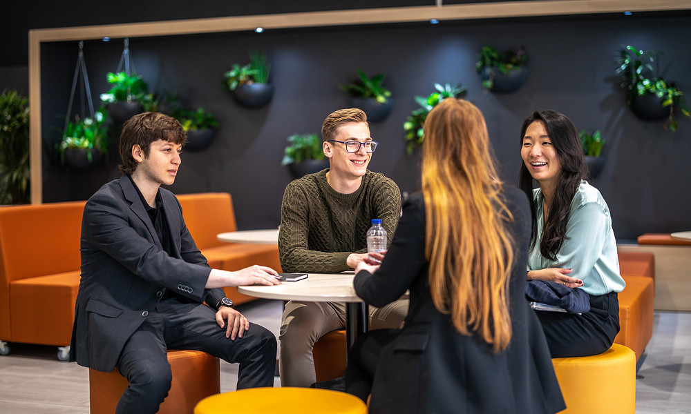 Four students sitting at table talking, in front of plant wall