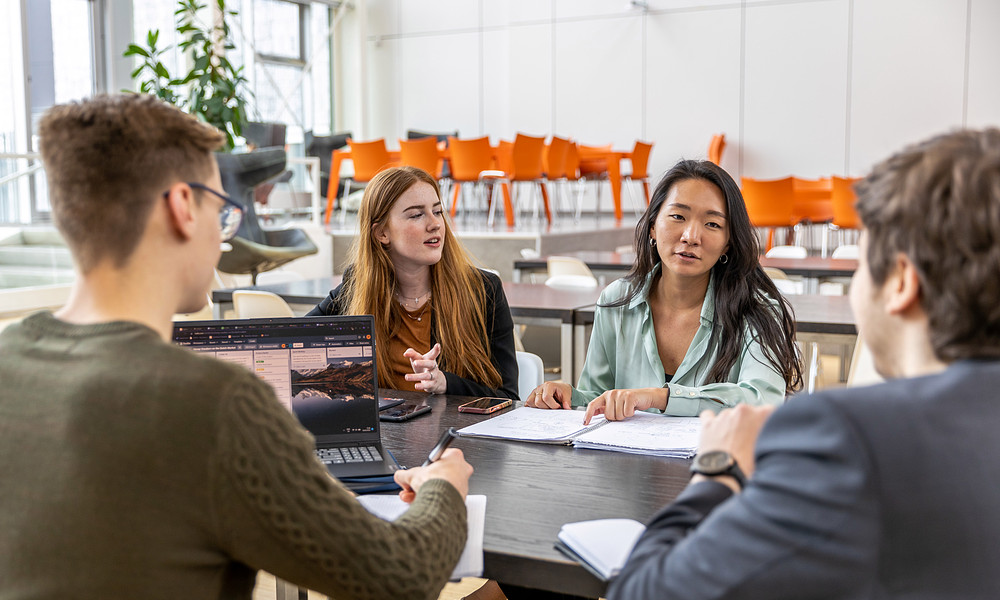 Four students discussing research at table with laptop and notebooks