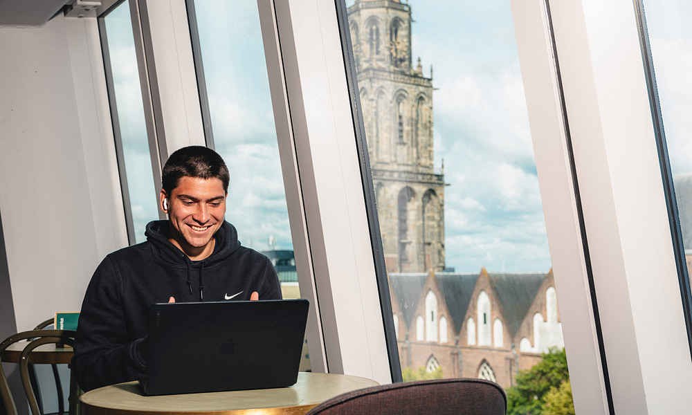 Student smiling while studying on a laptop in a modern building with large slanted windows, with the Martini Tower of Groningen visible in the background.