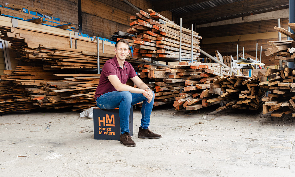 Alumnus Koen Rooseboom sitting on Master Up box at work with pile of wood boards behind him