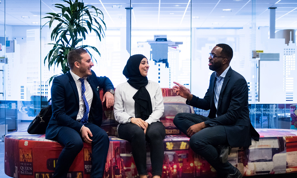 Three students from different backgrounds in business attire, talking on sofa.