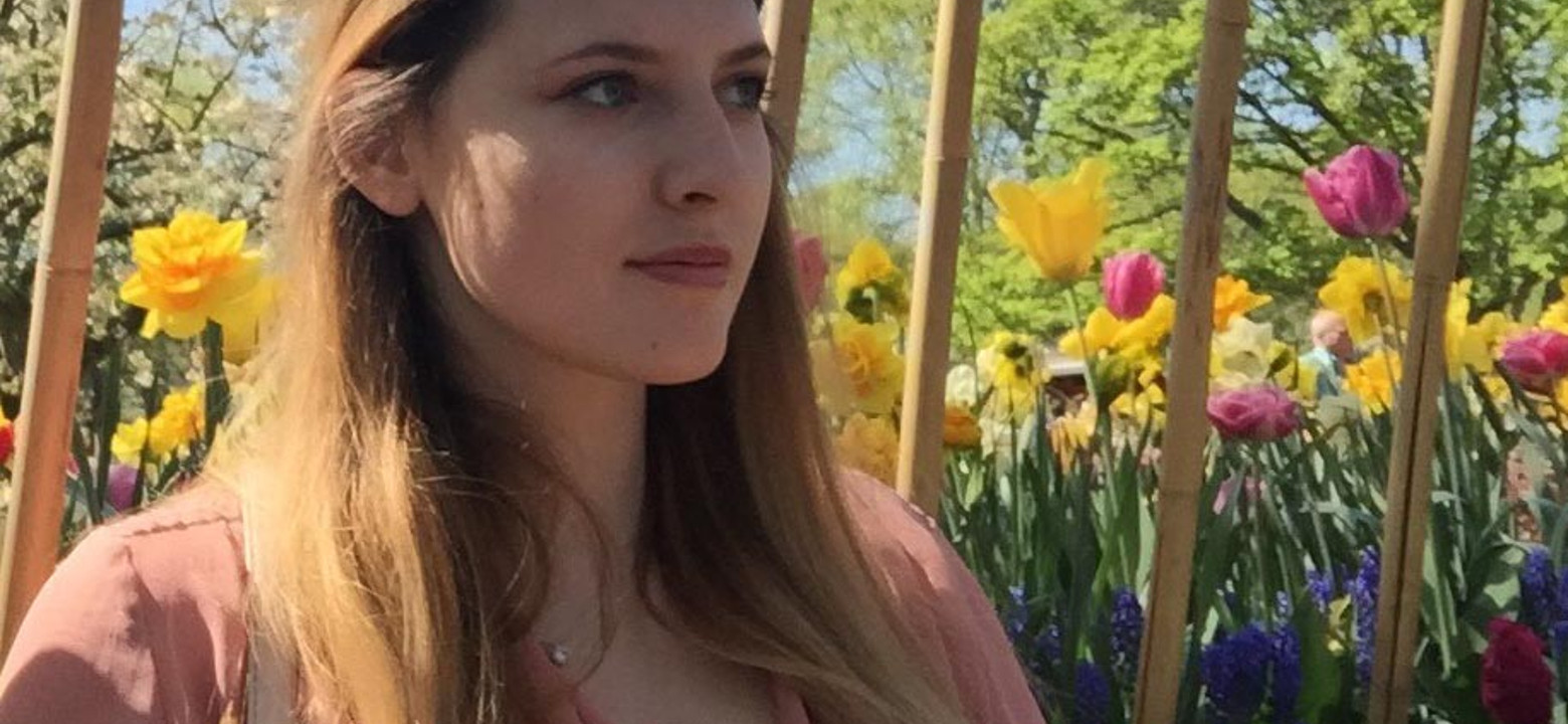 Young Romanian woman with long dark blonde hair looks off camera to the right. Behind her are trees and tulips.