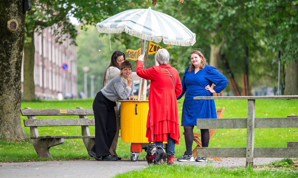 Drie vrouwen van het makerscollectief van de MensPlek staan met een gele kar in een park. Ze zijn in gesprek met een bejaarde dame.