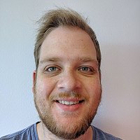 Een jonge man met blond haar, een snor en baard en een lichtblauw t-shirt kijkt glimlachend in de camera.