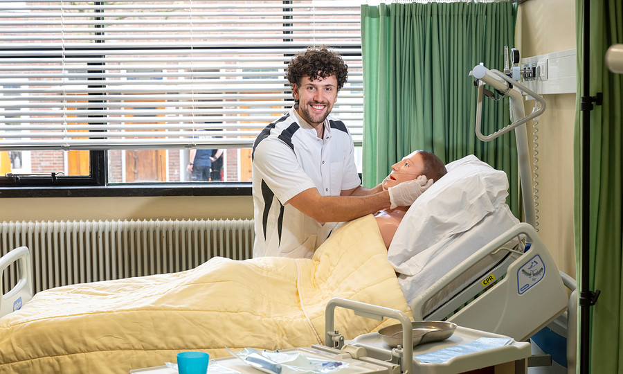 Verpleegkundige kijkt in camera en doet handeling bij oefenpop in verpleegbed