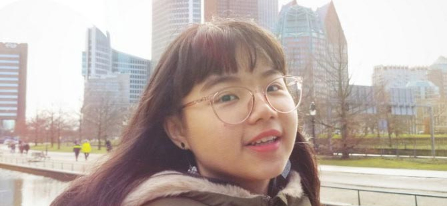 Young Vietnamese woman with long dark hair and glasses looks into the camera. She is wearing a green parka. Behind her you can see the city of Enschede.
