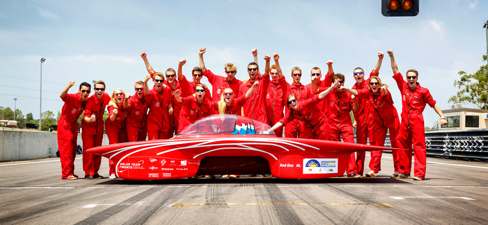 Teamfoto van Solar Team Twente. De teamleden staan achter de rode racewagen en steken hun hand op.