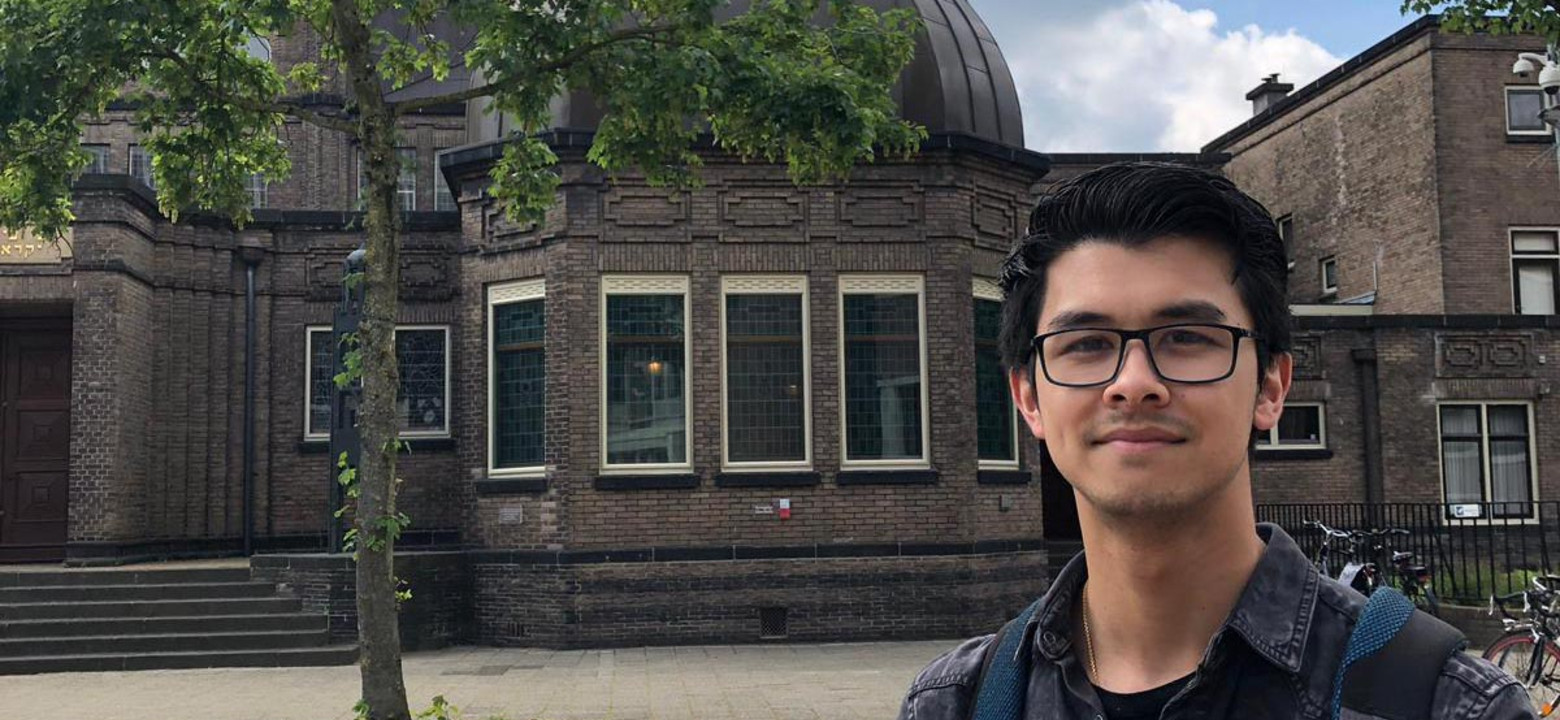 Young German man with short dark hair and glasses looks into the camera. He is wearing a jean jacket and a backpack. Behind him is a street with a tree and historial looking building.